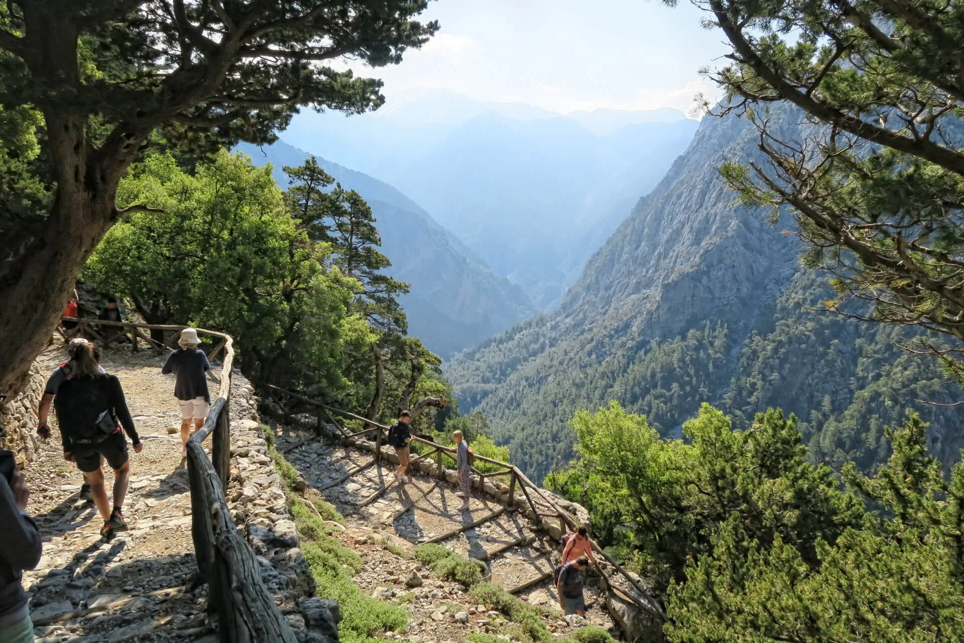 Best travel agency in Greece Les randonneurs en petits groupes dans le nord de la Grèce marchent le long d'un sentier de montagne clôturé avec des arbres luxuriants et des sommets montagneux lointains sous un ciel clair en arrière-plan. Offbeat-Travel