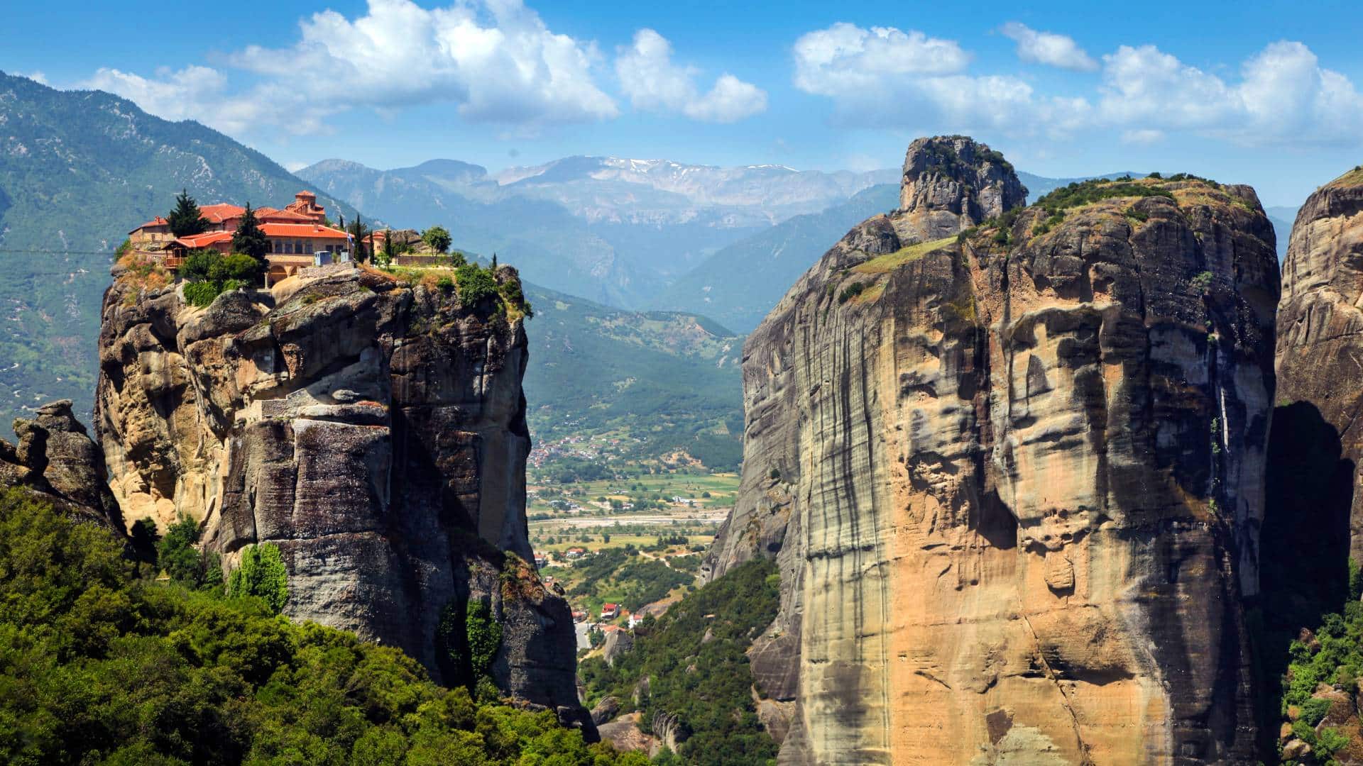 Best travel agency in Greece A monastery sits atop a rocky cliff in Meteora Greece, embraced by lush greenery and rugged mountains under a partly cloudy sky, offering unparalleled views for those on hiking tours in Meteora. Offbeat-Travel