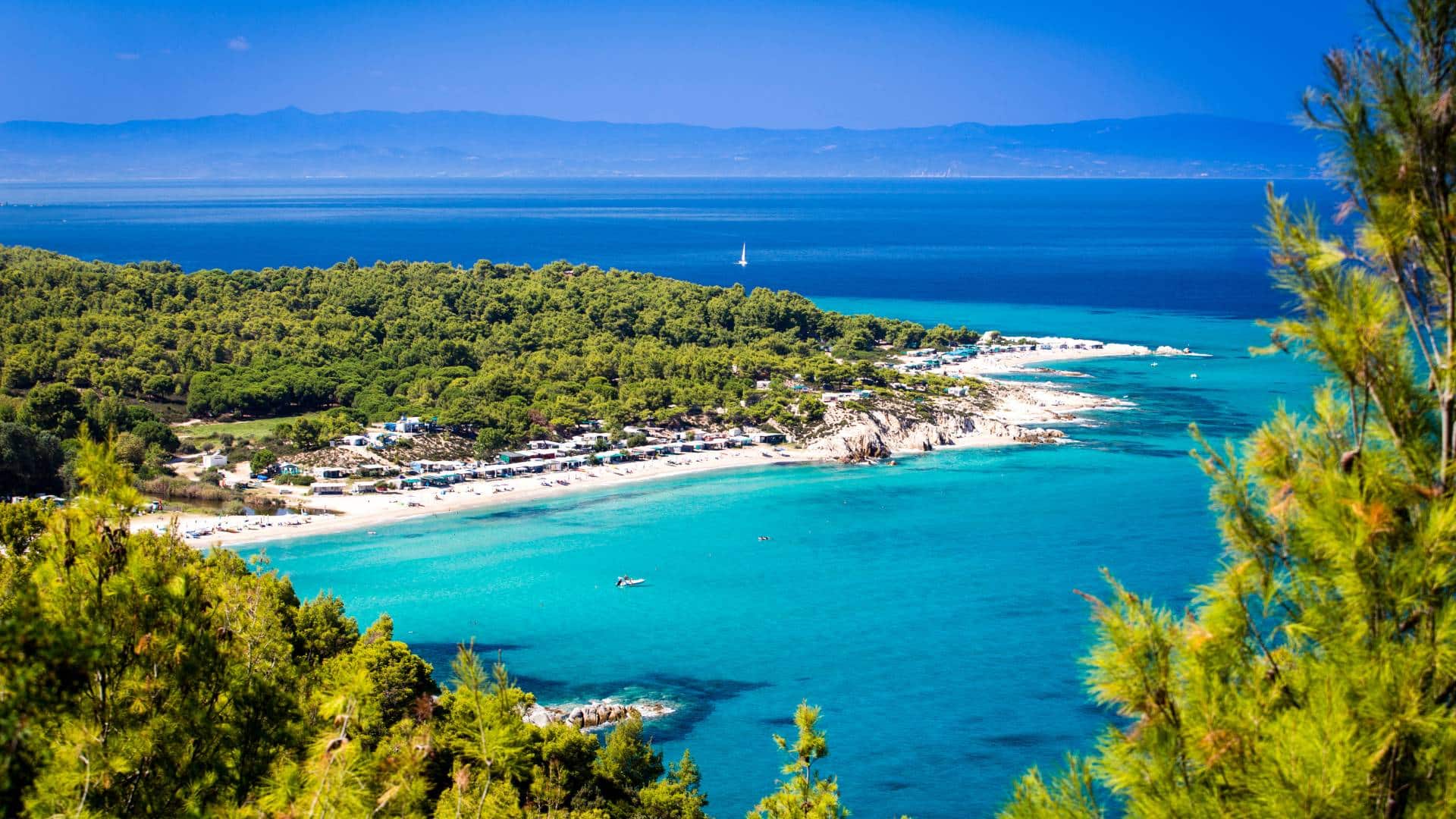 Best travel agency in Greece A scenic coastline in Chalkidiki Greece in Sithonia, boasts turquoise waters, a sandy beach, and lush green trees. A small boat gently floats while distant mountains rise under the clear blue sky. This picturesque vista captures the serene beauty of Halkidiki beaches. Offbeat-Travel