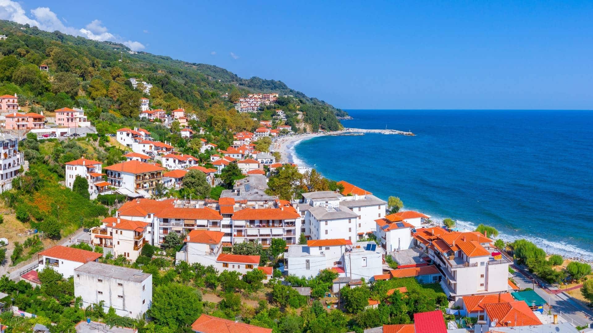 Organise your trip to Greece. Best travel agency in Greece Aerial view of a coastal village with red-roofed buildings, surrounded by green hills and a blue sea under a clear sky—a perfect stop on your road trip through Greece. Offbeat-Travel